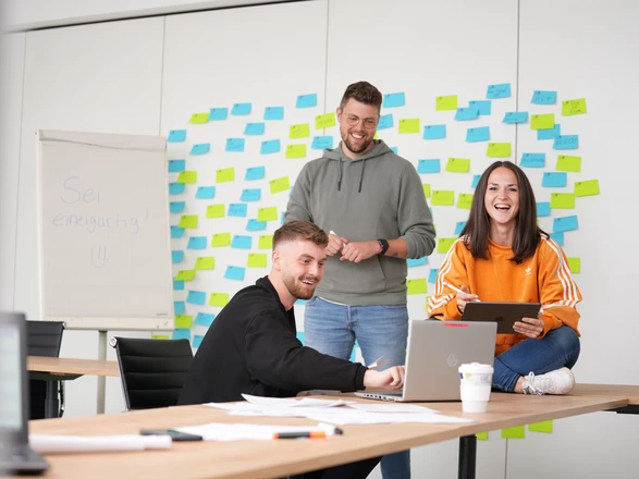 Zwei junge Männer und eine junge Frau arbeiten kreativ zusammen vor einem Laptop. Im Hintergrund hängen viele Papier-Karten an der Wand.